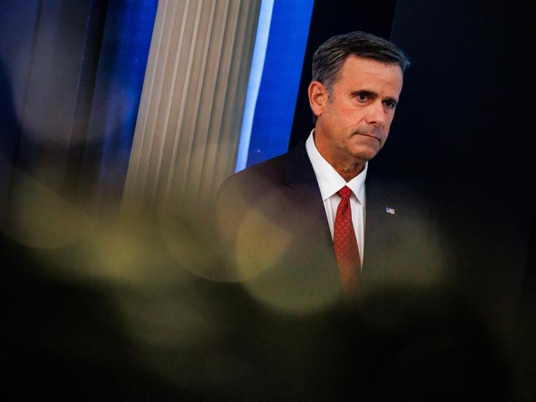 John Ratcliffe trägt eine schwarze Anzugjacke, ein weißes Hemd und eine rote Krawatte und steht vor einem dunklen Hintergrund. Sein Blick geht zur Seite.