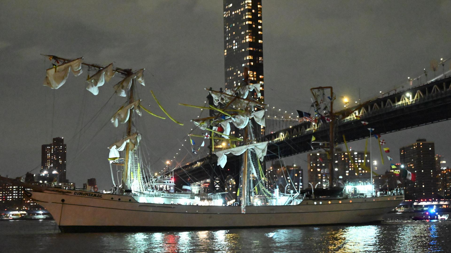 Das Segel-Schiff ist halb unter der Brücke in der Stadt New York im Land USA. Die Masten an dem Schiff sind umgeknickt. Es ist Nacht. Das Schiff ist hell beleuchtet. Im Hintergrund sind die Hoch-Häuser von New York. Das Segel-Schiff ist halb unter der Brücke in der Stadt New York im Land USA. Die Masten an dem Schiff sind umgeknickt. Es ist Nacht. Das Schiff ist hell beleuchtet. Im Hintergrund sind die Hoch-Häuser von New York.