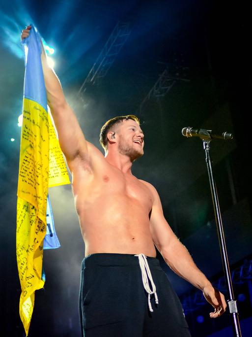 Frontmann Dan Reynolds steht halbnackt auf einer erleuchteten Bühne und hält eine Fahne der Ukraine hoch und animiert seine Fans.