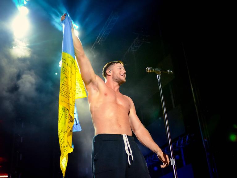 Frontmann Dan Reynolds steht halbnackt auf einer erleuchteten Bühne und hält eine Fahne der Ukraine hoch und animiert seine Fans.