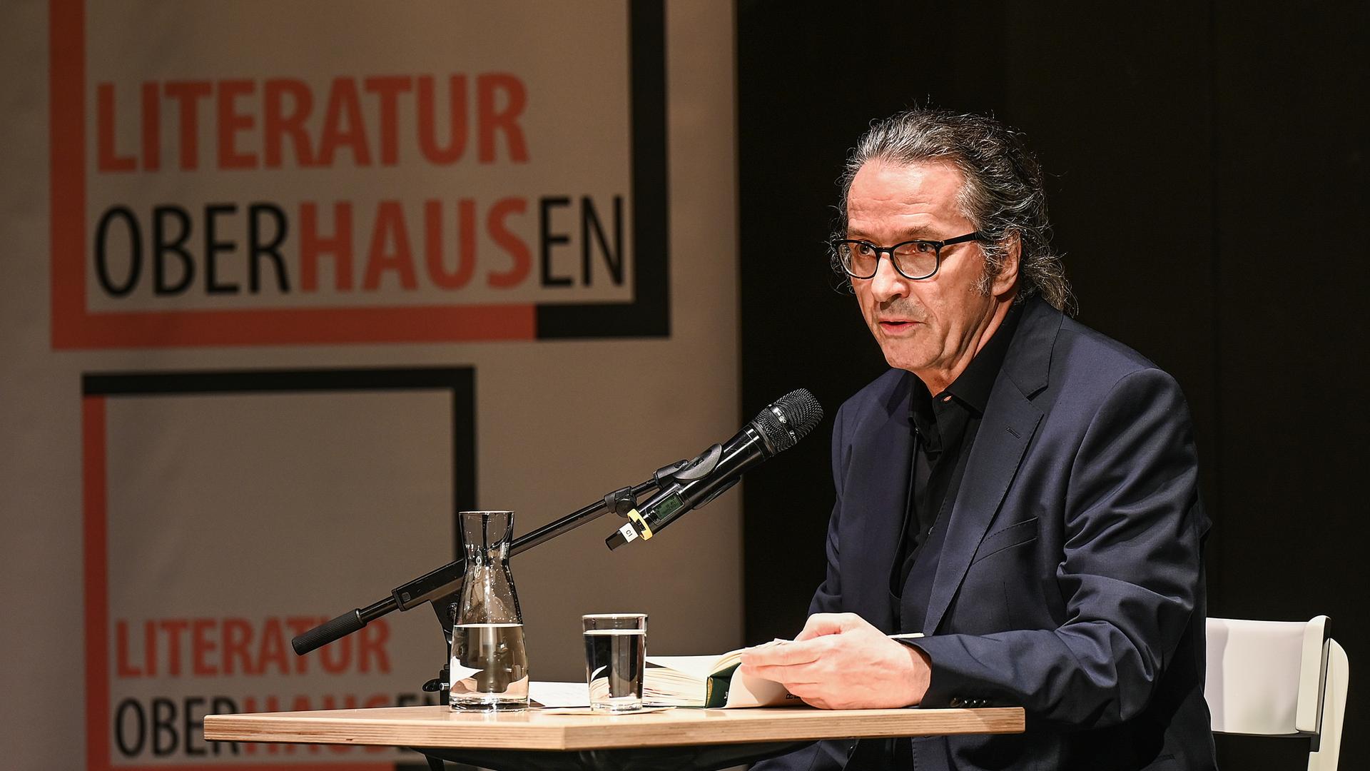 Der Schriftsteller Ralf Rothmann bei einer Lesung in Oberhausen. Er sitzt an einem Tisch im schwarzen Anzug. Der Schriftsteller Ralf Rothmann bei einer Lesung in Oberhausen. Er sitzt an einem Tisch im schwarzen Anzug.
