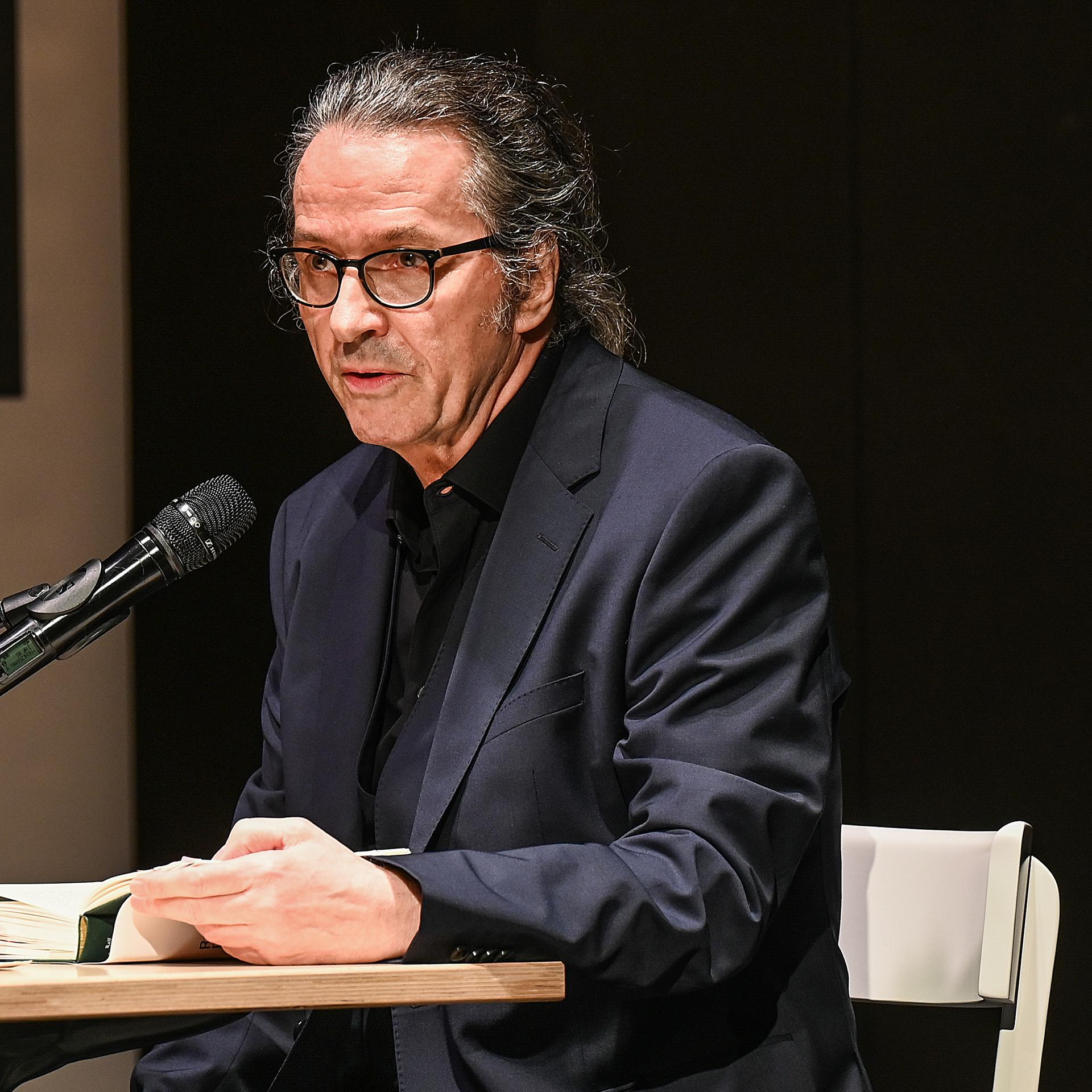 Der Schriftsteller Ralf Rothmann bei einer Lesung in Oberhausen. Er sitzt an einem Tisch im schwarzen Anzug. 