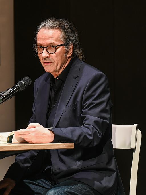 Der Schriftsteller Ralf Rothmann bei einer Lesung in Oberhausen. Er sitzt an einem Tisch im schwarzen Anzug. 
