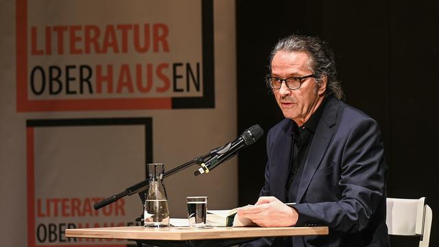 Der Schriftsteller Ralf Rothmann bei einer Lesung in Oberhausen. Er sitzt an einem Tisch im schwarzen Anzug. 