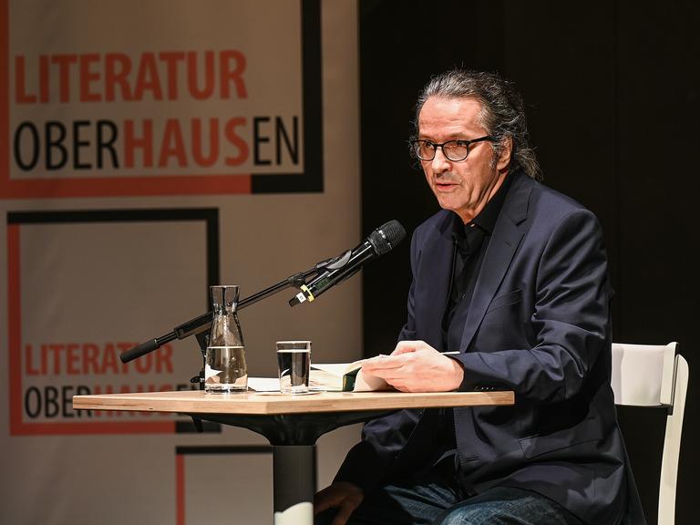 Der Schriftsteller Ralf Rothmann bei einer Lesung in Oberhausen. Er sitzt an einem Tisch im schwarzen Anzug. 