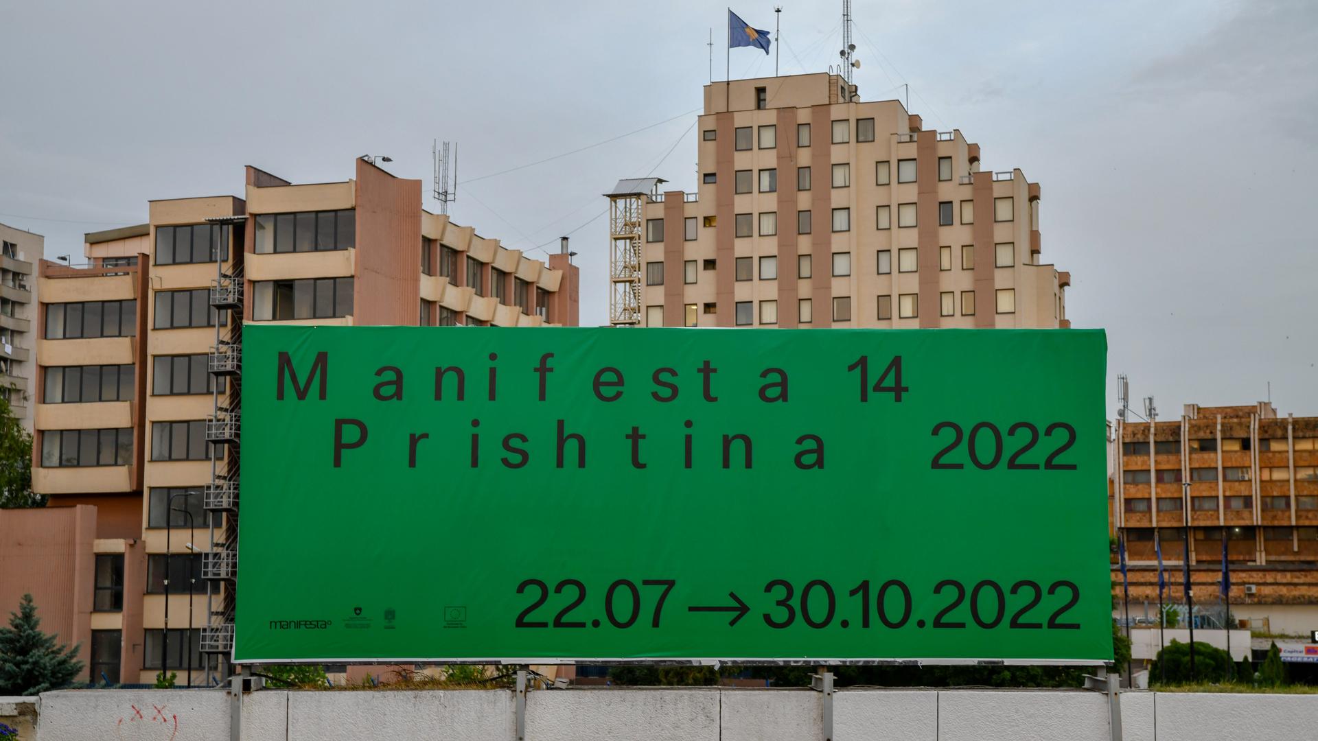 Ein großes günes Plakat mit der Aufschrift "Manifesta 14 / Prishtina 2022" hängt vor einem Wohnblock.