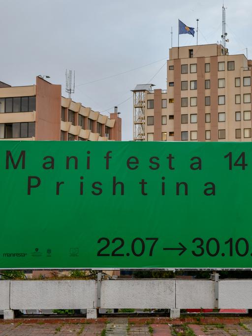 Ein großes günes Plakat mit der Aufschrift "Manifesta 14 / Prishtina 2022" hängt vor einem Wohnblock.