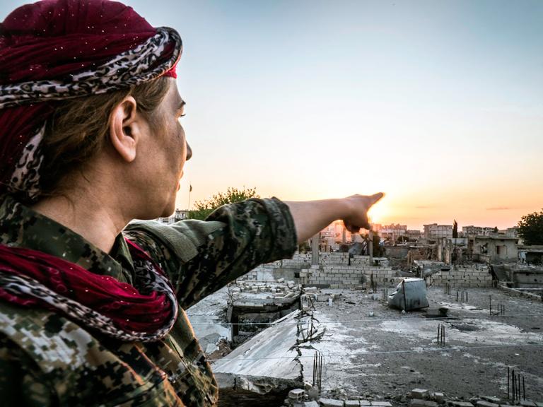 Eine kämpfende Frau in Rojava, Syrien mit Turban zeigt auf Häuser in der Ferne.