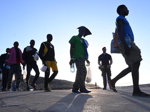 Flüchtlinge, die auf der Insel Lampedusa ankommen und in der Sonne laufen während sie von der Polizei beobachtet werden. 
