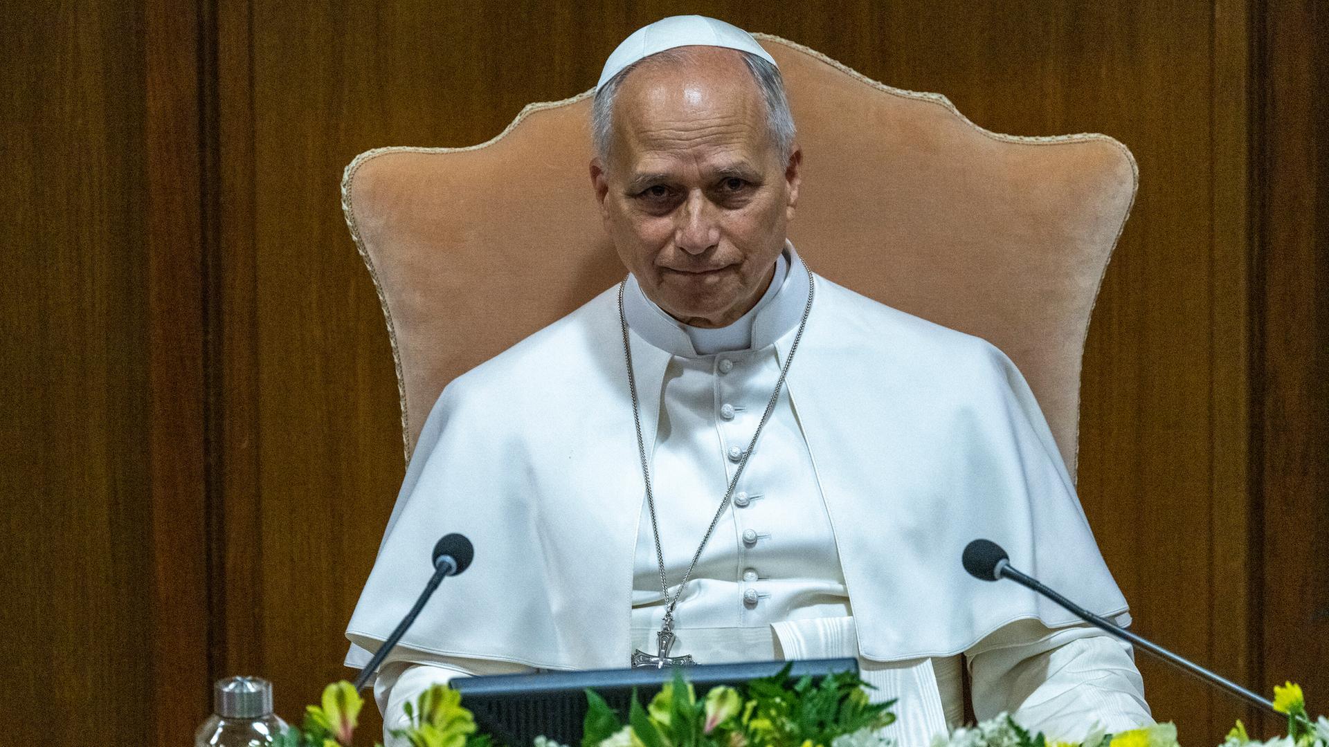 Papst Leo XIV. sitzt bei einer Pressekonferenz im Vatikan vor einem Mikrofon. Vor ihm schmücken Blumen das Rednerpult.