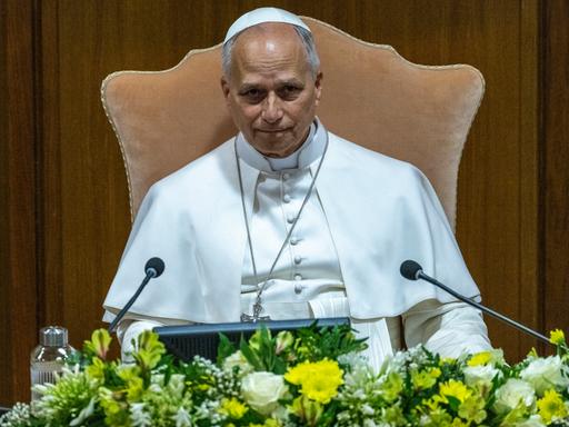 Papst Leo XIV. sitzt bei einer Pressekonferenz im Vatikan vor einem Mikrofon. Vor ihm schmücken Blumen das Rednerpult.