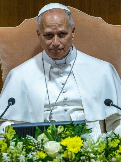 Papst Leo XIV. sitzt bei einer Pressekonferenz im Vatikan vor einem Mikrofon. Vor ihm schmücken Blumen das Rednerpult.