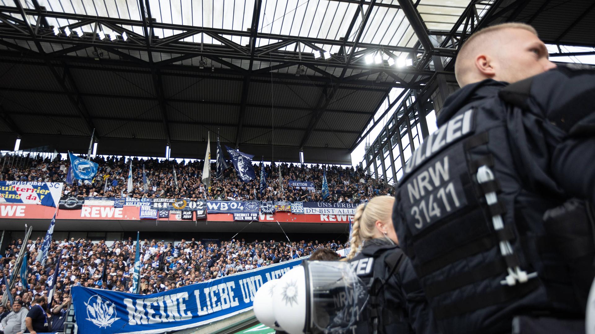 Ein Polizist steht mit dem Rücken zur Kamera am rechten Bildrand. Im Hintergrund sind Fans des Karlsruher SC auf einer Tribüne zu sehen.