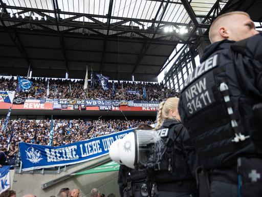 Ein Polizist steht mit dem Rücken zur Kamera am rechten Bildrand. Im Hintergrund sind Fans des Karlsruher SC auf einer Tribüne zu sehen.