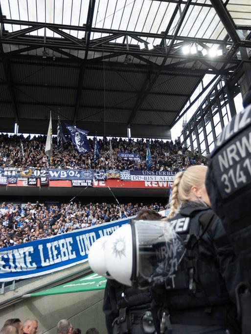 Ein Polizist steht mit dem Rücken zur Kamera am rechten Bildrand. Im Hintergrund sind Fans des Karlsruher SC auf einer Tribüne zu sehen.