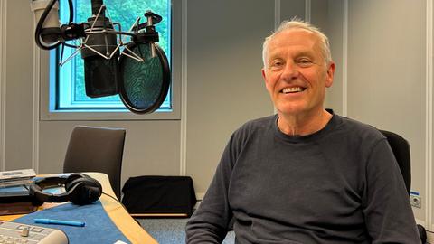 Ein freundlich lächelnder, grauhaariger Mann mit dunkelgrauem Pulli im Radiostudio mit Mikrophon an der Seite.