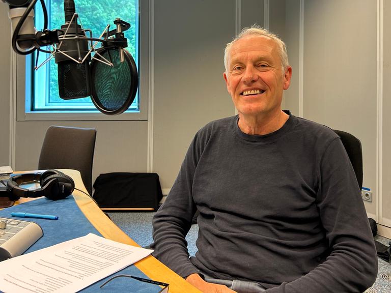 Ein freundlich lächelnder, grauhaariger Mann mit dunkelgrauem Pulli im Radiostudio mit Mikrophon an der Seite.