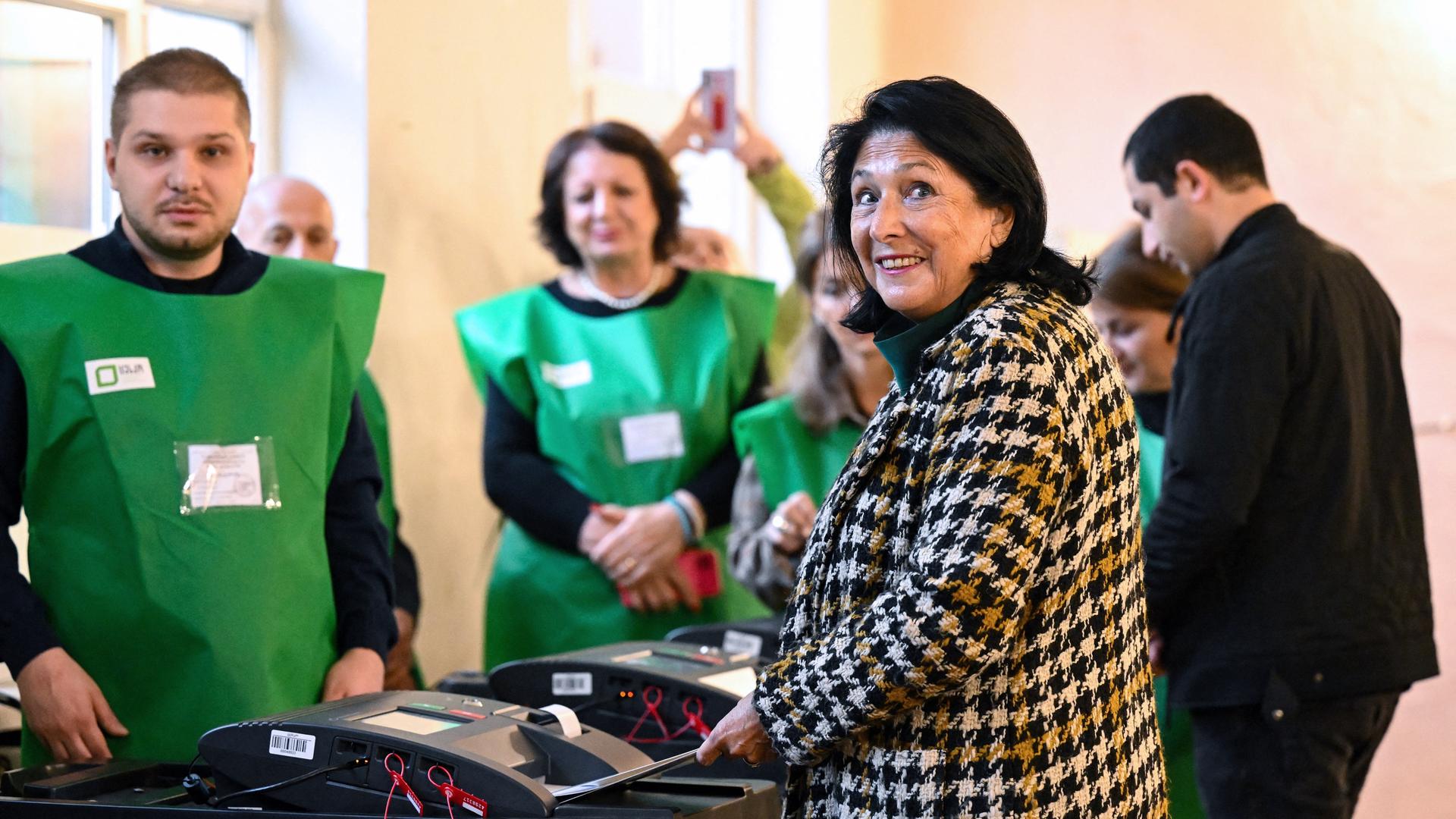 Eine Frau steckt ihren Wahlzettel in die Urne und schaut dabei in die Kamera, vor ihr stehen zwei Wahlhelfer in grünen Westen