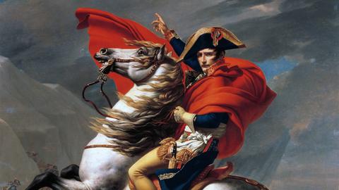 Das historische Ölgemälde zeigt den französischen Kaiser Napoleon Bonaparte in einer Uniform auf einem sich aufbäumenden Pferd sitzen. Mit der linken Hand hält er die Zügel, mit der rechten zeigt er nach oben.