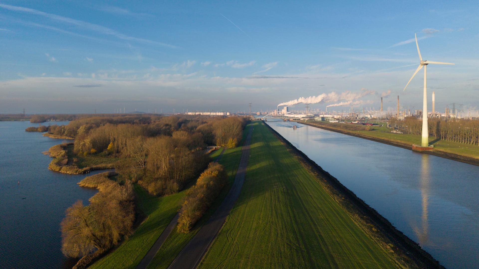 Eine Windturbine steht neben einem Kanal. Im Hintergrund sieht man Schornsteine und Rauch.