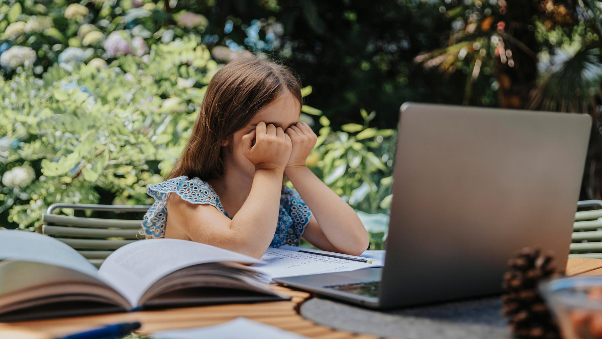 Ein Mädchen sitzt am Gartentisch und hält ihre Hände vor dem Gesicht. Vor ihr auf dem Tisch liegen Bücher, Hefte und ein Laptop.