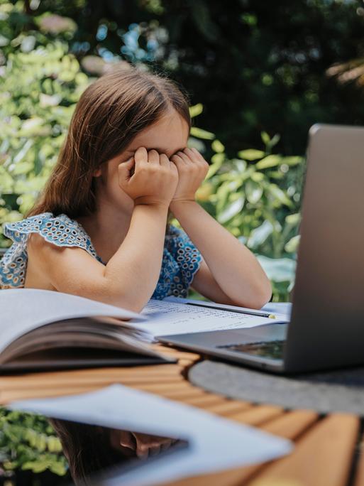 Ein Mädchen sitzt am Gartentisch und hält ihre Hände vor dem Gesicht. Vor ihr auf dem Tisch liegen Bücher, Hefte und ein Laptop.