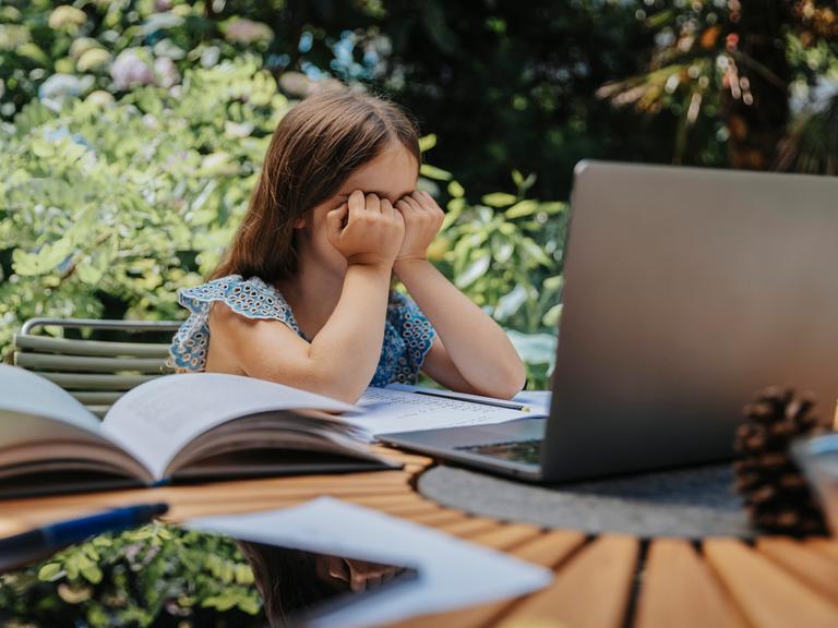 Ein Mädchen sitzt am Gartentisch und hält ihre Hände vor dem Gesicht. Vor ihr auf dem Tisch liegen Bücher, Hefte und ein Laptop.