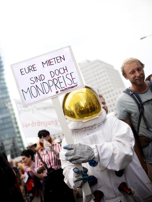 Ein als Astronaut verkleideter Mensch nimmt an einer Demo gegen zu hohe Mieten teil und trägt ein Schild, auf dem steht: "Eure Mieten sind doch Mondpreise"