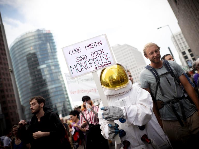 Ein als Astronaut verkleideter Mensch nimmt an einer Demo gegen zu hohe Mieten teil und trägt ein Schild, auf dem steht: "Eure Mieten sind doch Mondpreise"
