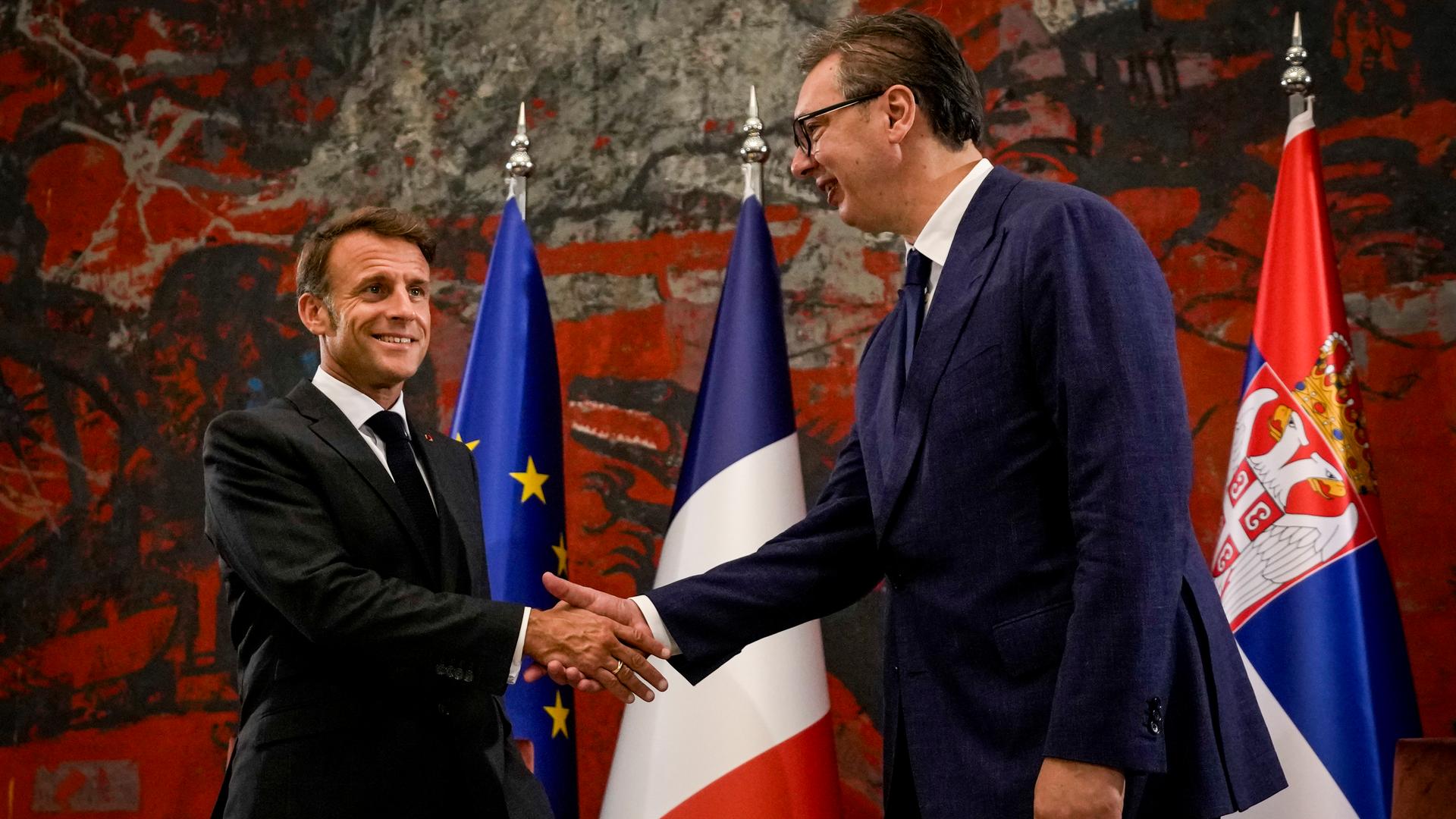 Der französische Präsident Macron und Serbiens Präsident Vucic schütteln sich die Hände. Hinter ihnen stehen mehrere Fahnen.