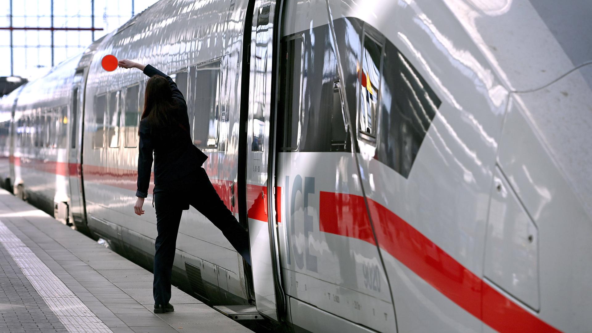 Eine Zugbegleiterin ist von hinten zu sehen, wie sie mit einem Bein auf dem Bahnsteig,  mit dem anderen Bein schon im Zug steht. Sie hält eine Kelle in die Luft.