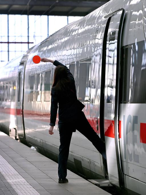 Eine Zugbegleiterin ist von hinten zu sehen, wie sie mit einem Bein auf dem Bahnsteig, mit dem anderen Bein schon im Zug steht. Sie hält eine Kelle in die Luft. Eine Zugbegleiterin ist von hinten zu sehen, wie sie mit einem Bein auf dem Bahnsteig, mit dem anderen Bein schon im Zug steht. Sie hält eine Kelle in die Luft.