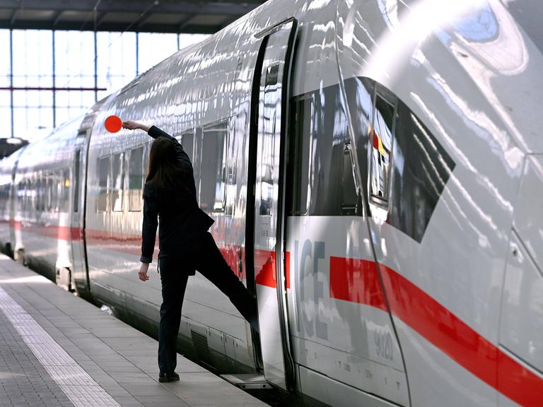 Eine Zugbegleiterin ist von hinten zu sehen, wie sie mit einem Bein auf dem Bahnsteig, mit dem anderen Bein schon im Zug steht. Sie hält eine Kelle in die Luft. Eine Zugbegleiterin ist von hinten zu sehen, wie sie mit einem Bein auf dem Bahnsteig, mit dem anderen Bein schon im Zug steht. Sie hält eine Kelle in die Luft.