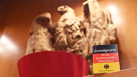 Vor dem Bundesadler im Sitzungssaal des Bundesverfassungsgericht steht eine Miniaturausgabe des Grundgesetz für die Bundesrepublik Deutschland sowie ein Barett eines Bundesverfassungsrichters. 