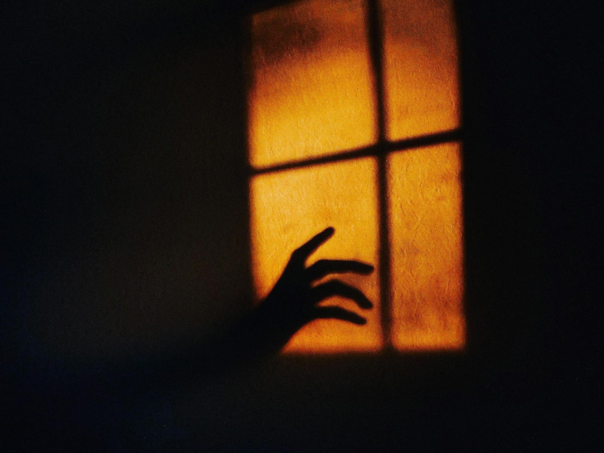 Auf einer Wand erscheint das gelbe Licht eines Fensters, in das der Schatten einer Hand hineinragt.
