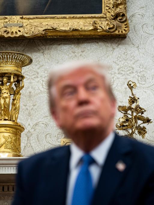 US-Präsident Trump sitzt im Oval Office des Weißen Hauses. Hinter ihm stehen ein goldener Pokal und weitere goldene Dekorationsobjekte. 