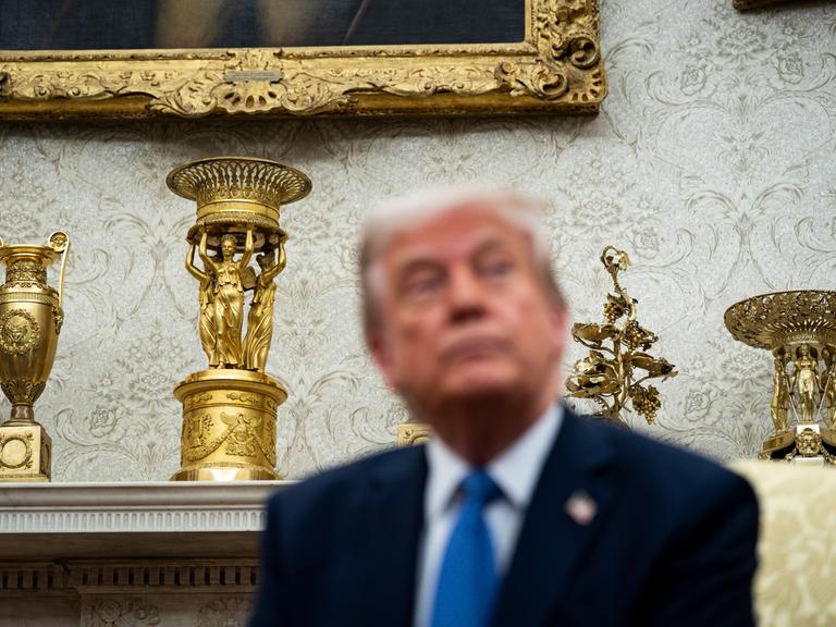 US-Präsident Trump sitzt im Oval Office des Weißen Hauses. Hinter ihm stehen ein goldener Pokal und weitere goldene Dekorationsobjekte. 