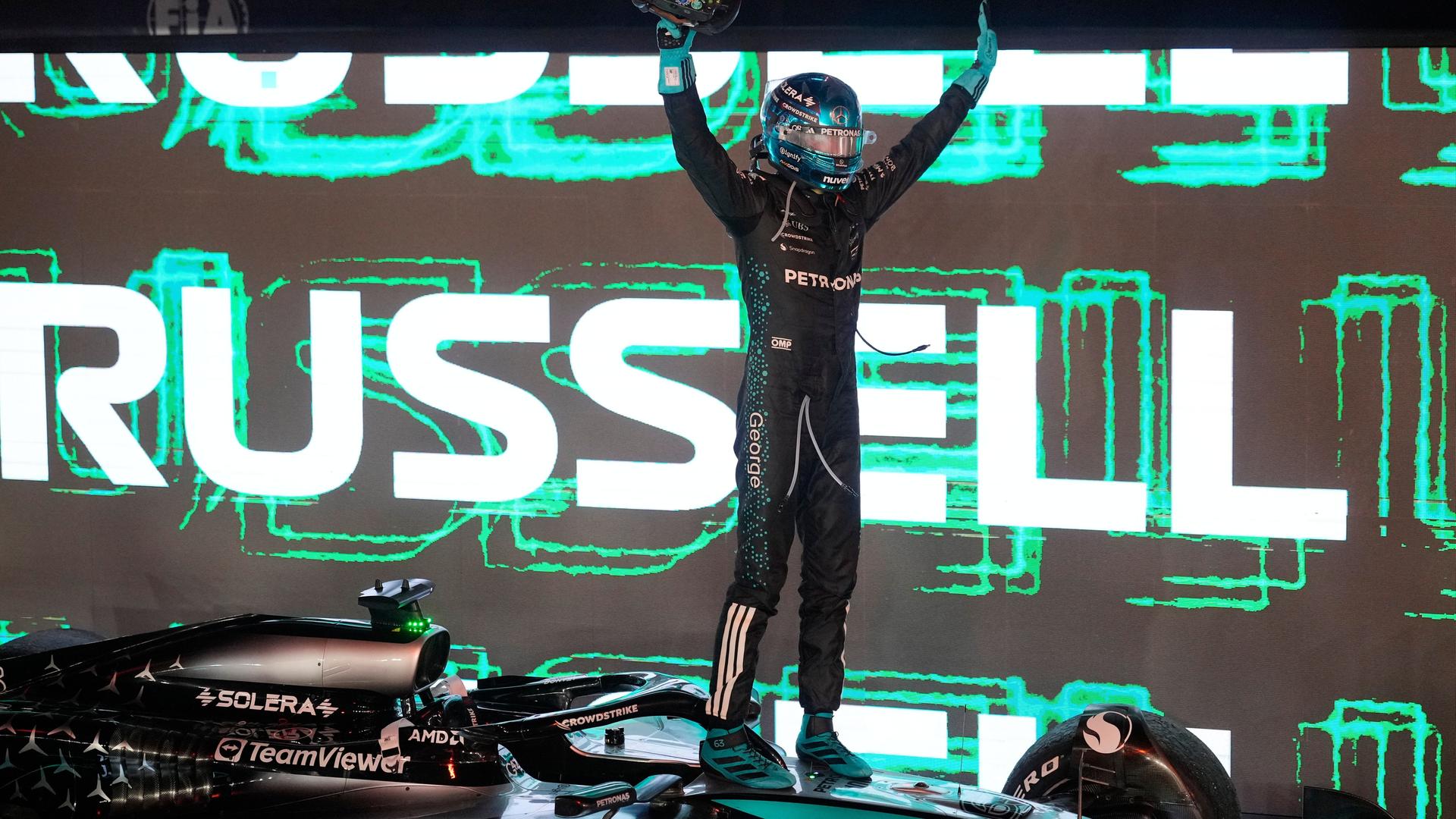 Das Bild zeigt George Russell mit Helm und Rennanzug. Er steht auf seinem Formel-1-Rennwagen und jubelt. Im Hintergrund ist sein Name auf einem großen Display zu sehen.