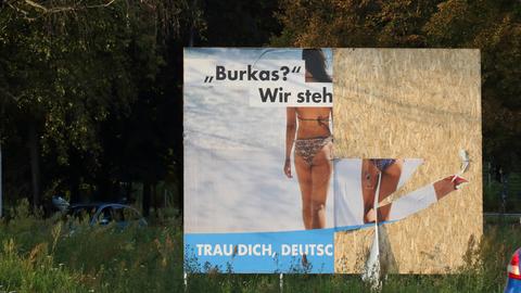 Teilweise zerstörtes Wahlplakat der AfD, auf dem zwei Frauen von hinten zu sehen sind. Sie tragen beide Bikinis. Die Aufschrift beginnt mit "Burkas? Wir steh". Weiter ist es nicht zu lesen.