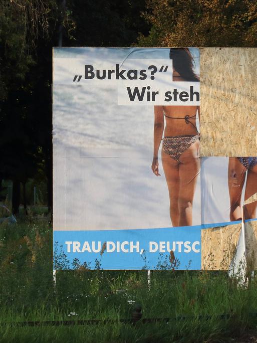 Teilweise zerstörtes Wahlplakat der AfD, auf dem zwei Frauen von hinten zu sehen sind. Sie tragen beide Bikinis. Die Aufschrift beginnt mit "Burkas? Wir steh". Weiter ist es nicht zu lesen.