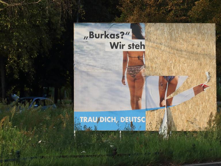 Teilweise zerstörtes Wahlplakat der AfD, auf dem zwei Frauen von hinten zu sehen sind. Sie tragen beide Bikinis. Die Aufschrift beginnt mit "Burkas? Wir steh". Weiter ist es nicht zu lesen.
