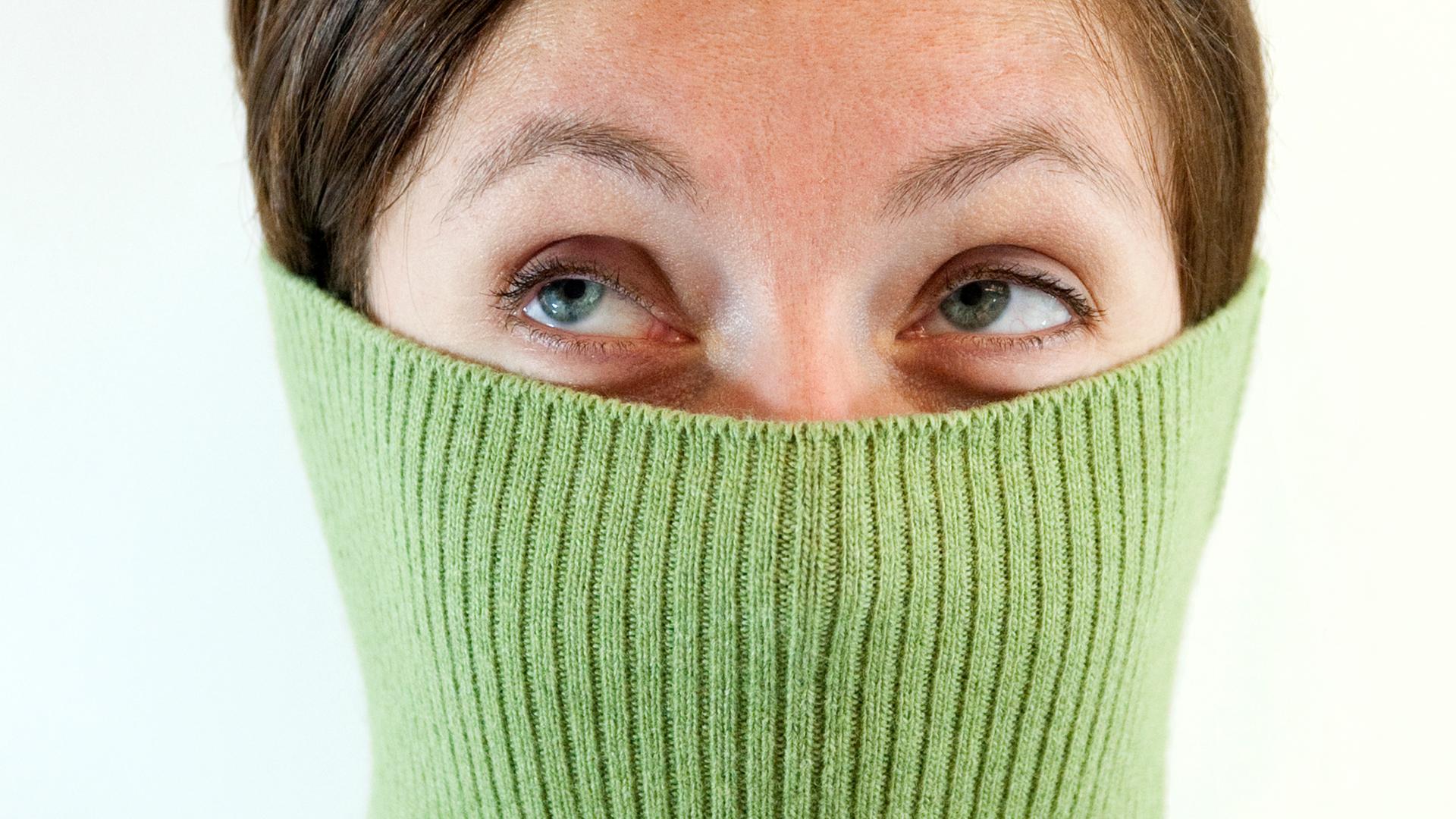 Eine Frau hat ihren Pullover bis über die Nase gezogen und schaut beschämt noch oben links. Ihr steigt Schamesröte ins Gesicht. 