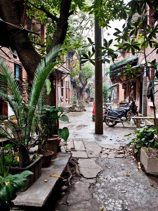 Blick in die Siedlung Tongyuanju in Chongqing, auf beiden Seiten eines schmalen Wegs rote Ziegelsteinhäuser, davor ein Motorrad und Pflanzen.