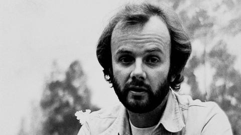 John Peel kniet auf dem Schwarz-Weiß-Foto im Freien. Der Hintergrund ist unscharf. Er trägt Bart und hat eine Jeans-Jacke und eine Jeans-Hose an.