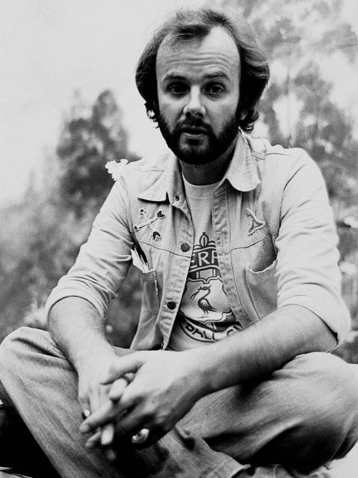 John Peel kniet auf dem Schwarz-Weiß-Foto im Freien. Der Hintergrund ist unscharf. Er trägt Bart und hat eine Jeans-Jacke und eine Jeans-Hose an.