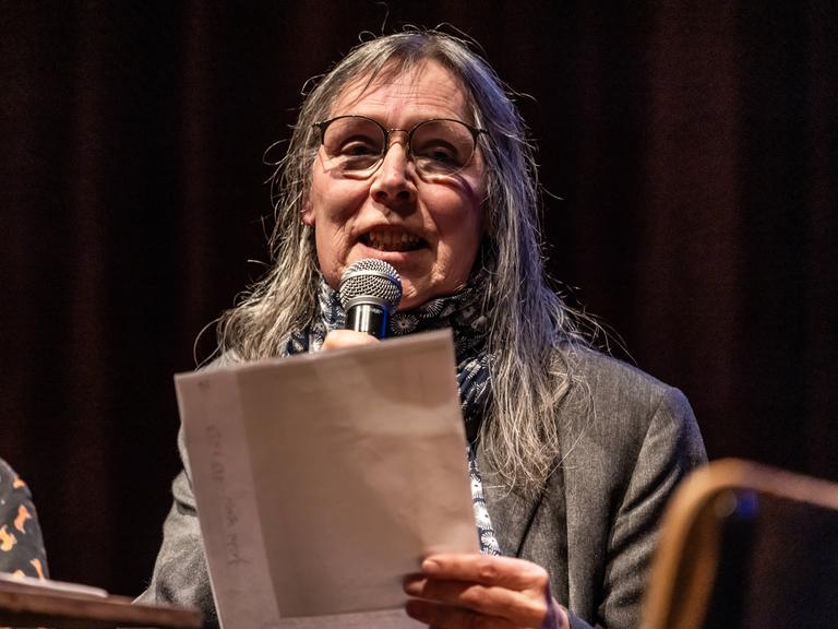 Jayne-Ann Igel liest in der Volksbühne am Rosa-Luxemburg-Platz. Sie sitzt vor einem Mikrofon und hält Zettel in der Hand.