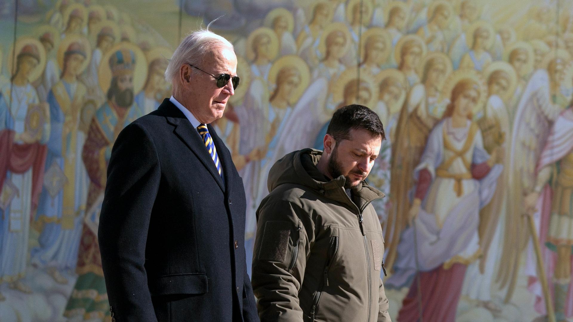 Joe Biden läuft bei einem Staatsbesuch in der Ukraine neben Wolodymyr Selenskyj vor einer Wand mit Heiligendarstellungen.