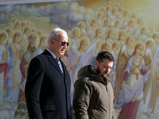 Joe Biden läuft bei einem Staatsbesuch in der Ukraine neben Wolodymyr Selenskyj vor einer Wand mit Heiligendarstellungen.