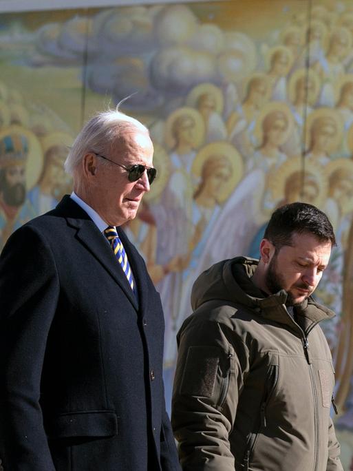 Joe Biden läuft bei einem Staatsbesuch in der Ukraine neben Wolodymyr Selenskyj vor einer Wand mit Heiligendarstellungen.