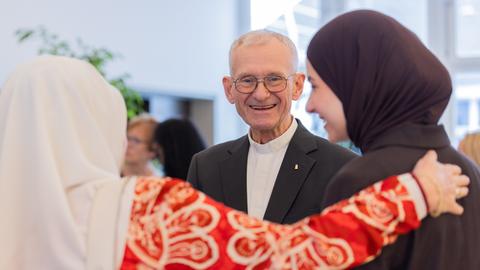 Pfarrer Franz Meurer unterhält sich mit Frauen, die Hidschabs tragen. Er trägt eine Brille und lächelt in die Kamera.